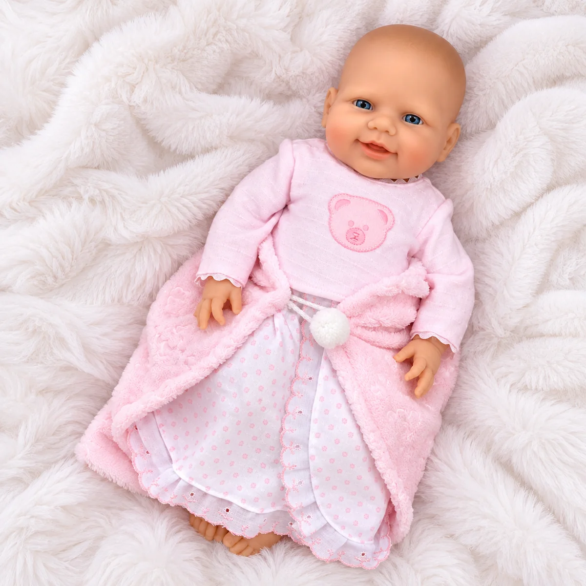 Bambolotto disteso su una coperta bianca a pelo lungo, copertina rosa aperta e pompon bianco ben visibile